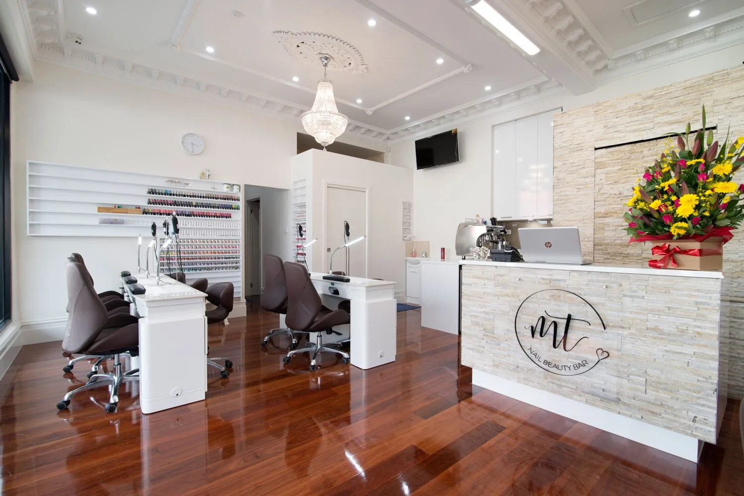 Wide interior view — manicure stations, the full polish wall, stacked-stone reception with the MT logo, crystal chandelier and glossy timber floor