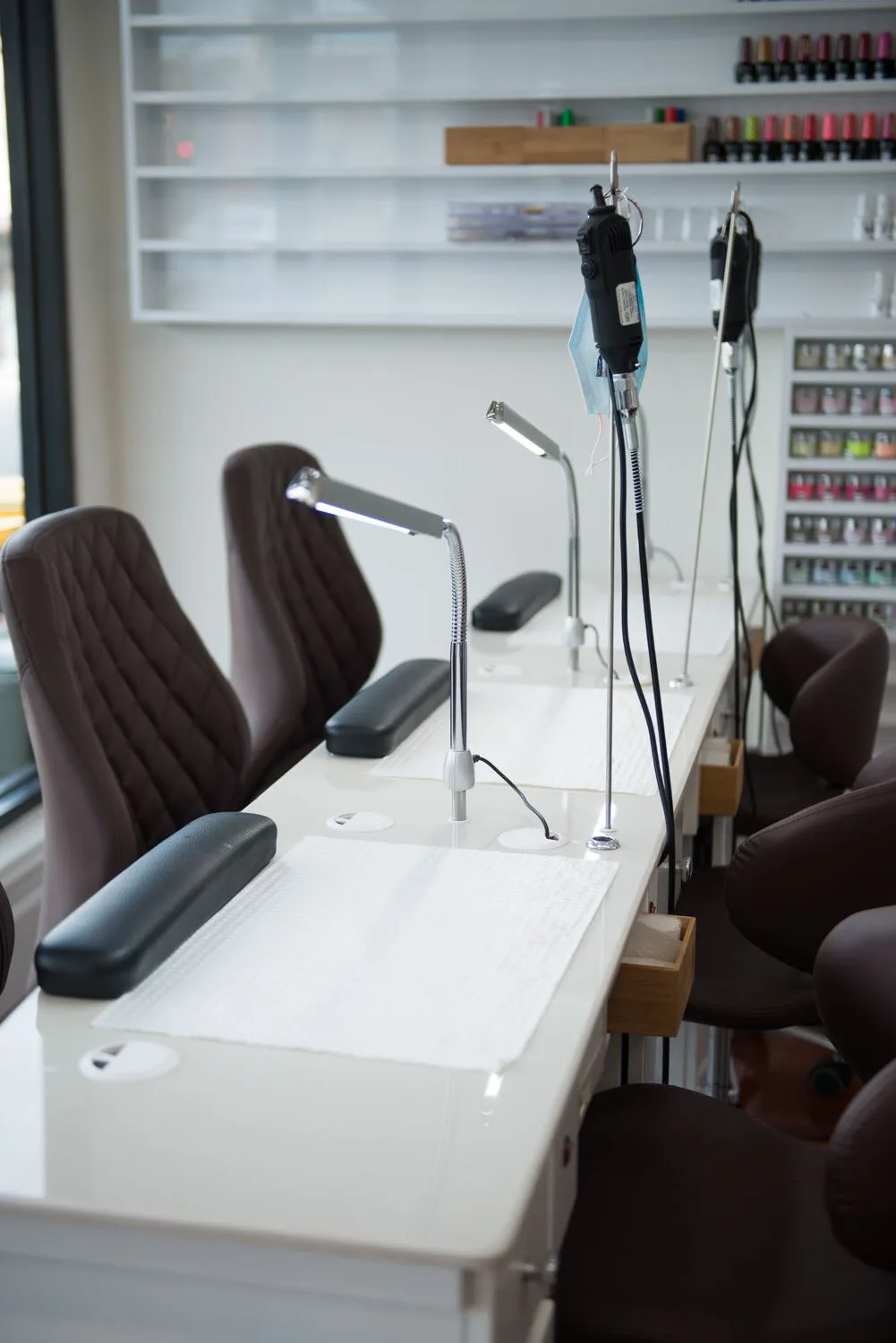 A row of manicure stations with drill lamps, and the floor-to-ceiling polish-bottle wall beside a window